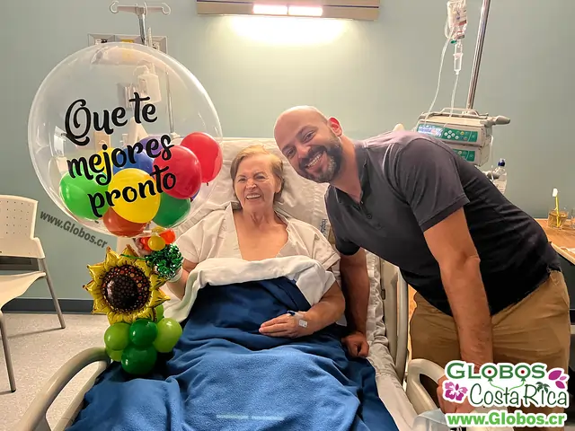 Señora sonriente en una cama de hospital sosteniendo un globo con la leyenda "Que te mejores pronto" y un detalle de girasol.