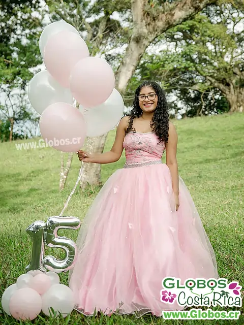 Quinceañera en un vestido rosa con globos blancos y rosados y decoración de número 15 en un jardín.