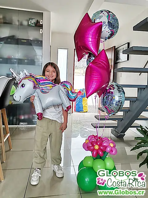 Niña sonriente con decoración de globos de unicornio y flores coloridas en una sala iluminada.